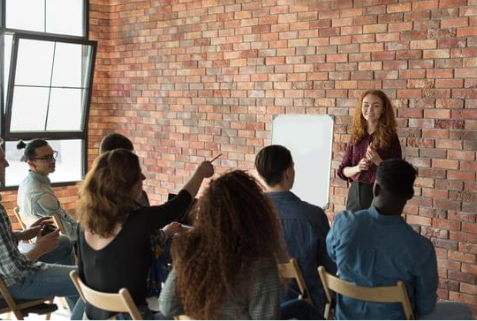 Photo of a group of people sitting in chairs facing a woman with red hair leading them in a discussion 